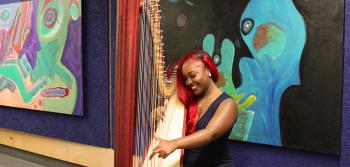 A smiling student plays the harp in front of colorful paintings.