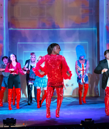 The image shows a scene from the musical "Kinky Boots," featuring performers in bright red knee-high boots. The central performer, in a red sequin dress, leads the group in a lively dance. The stage is vibrant, with colorful lighting and costumes, including a Union Jack dress and a suit.